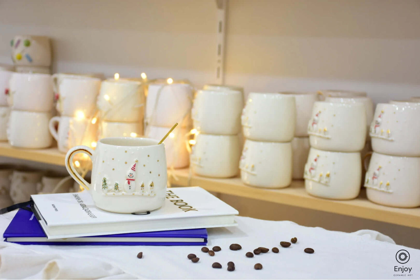 A festive ceramic mug with a snowman and gold-accented trees sits on stacked books, surrounded by scattered coffee beans, with cozy lights in the background.