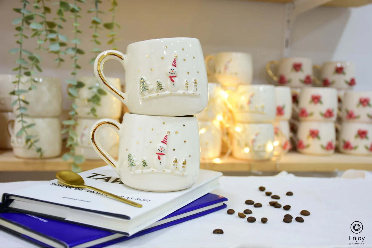 Two stacked ceramic mugs with festive snowman and tree designs, gold accents, and cozy background lights, set on books with scattered coffee beans.