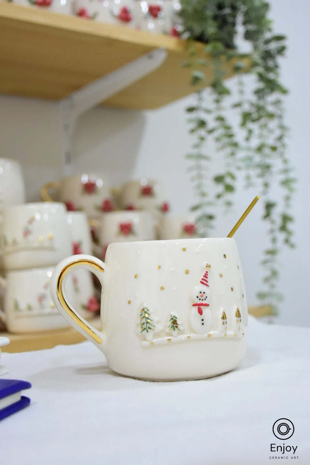 Festive ceramic mug featuring a snowman, small trees, and gold accents. Placed with a cozy backdrop of stacked mugs and hanging greenery.