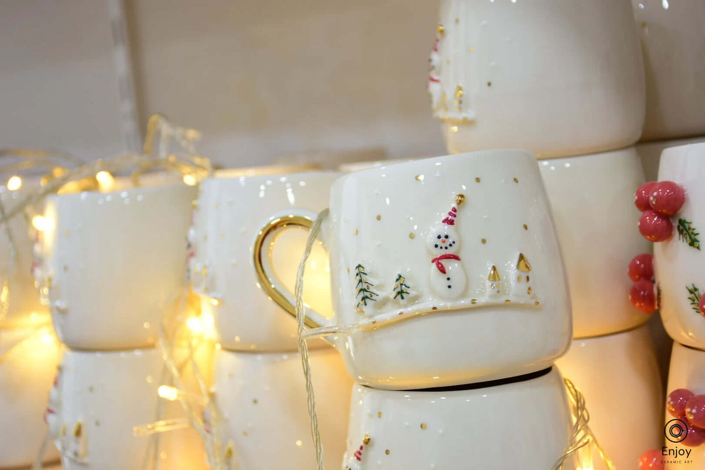 A holiday-themed ceramic mug with a snowman, trees, and gold accents, surrounded by warm fairy lights for a cozy festive ambiance.