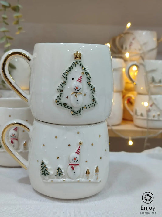 Two handmade ceramic mugs featuring festive 3D designs: a snowman framed in a Christmas tree and a snowy scene with a snowman, both with gold handles.