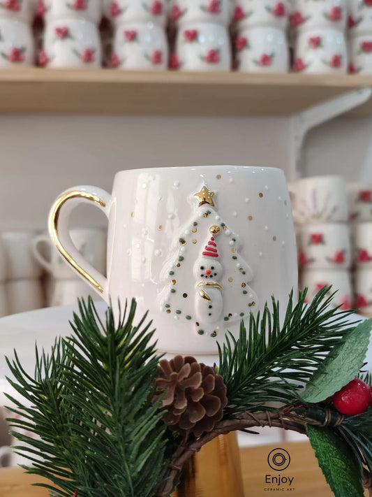 A handmade ceramic mug featuring a snowman framed by a Christmas tree, with golden accents and festive details. The mug is displayed on a white stand, surrounded by pine branches and pinecones.