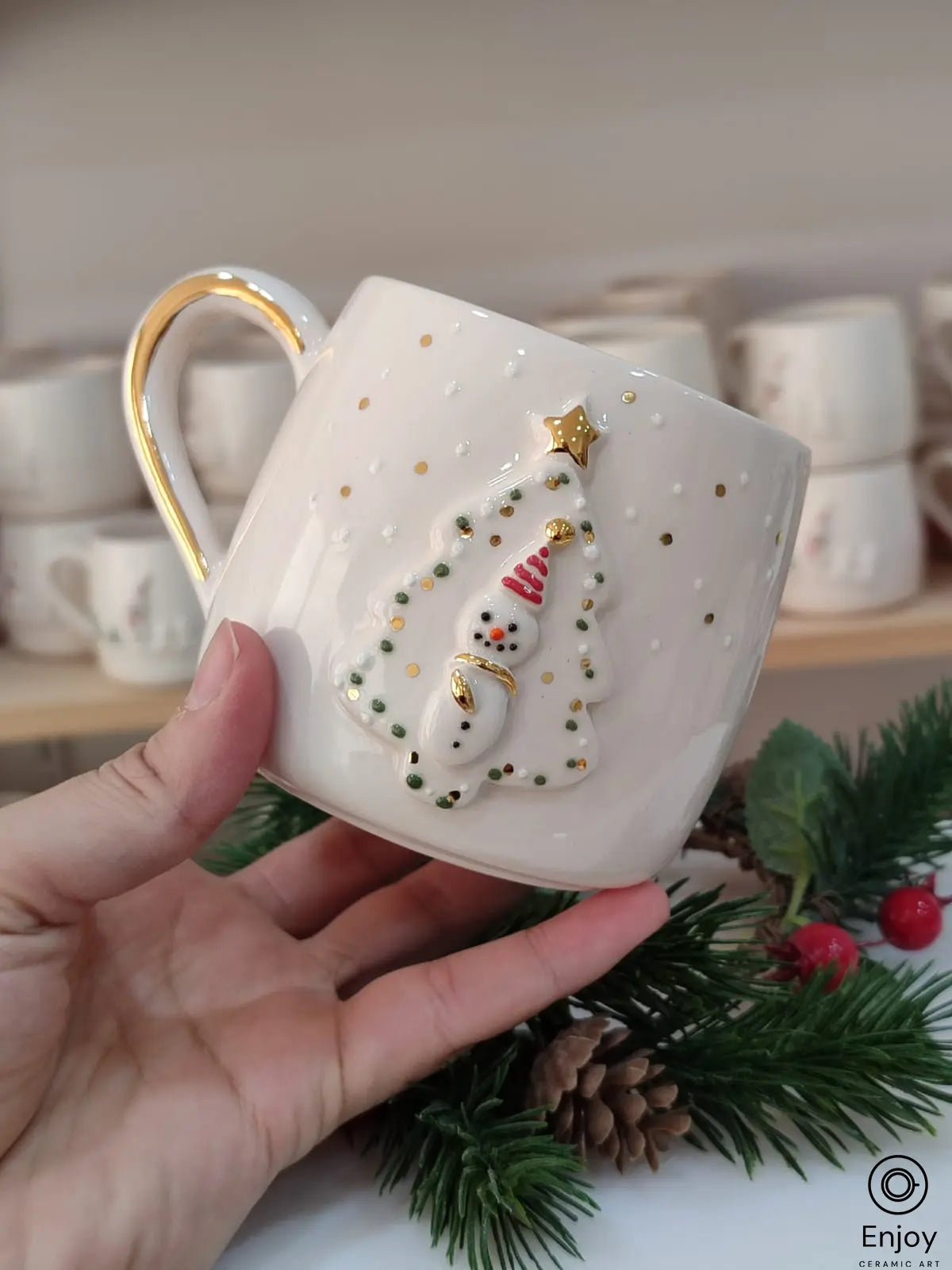 A festive handmade ceramic mug featuring a snowman framed by a Christmas tree with golden accents and snow-like detailing. It is being held up, with pine branches and berries in the background for a holiday display.