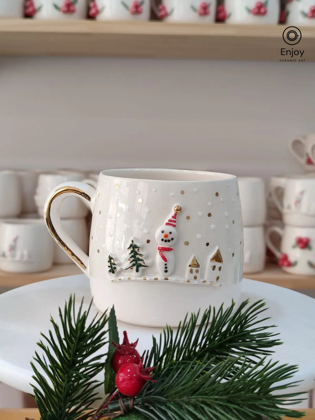 A festive handmade ceramic mug featuring a joyful snowman in a snowy village, with gold accents. It’s displayed on a white stand, adorned with pine branches and red berries.