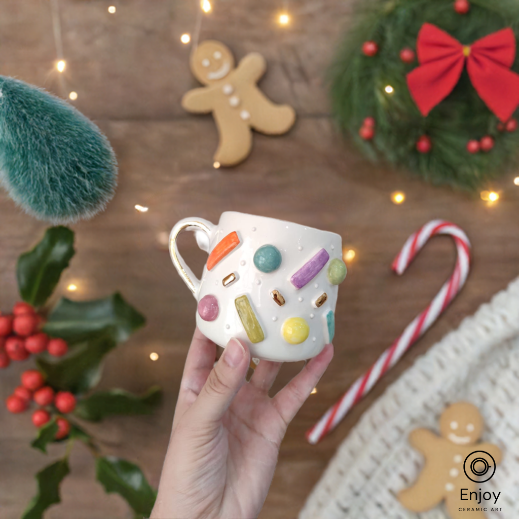 Hand holding a ceramic mug adorned with colorful sprinkles against a festive backdrop of holiday decor, including gingerbread, a candy cane, and a Christmas wreath.