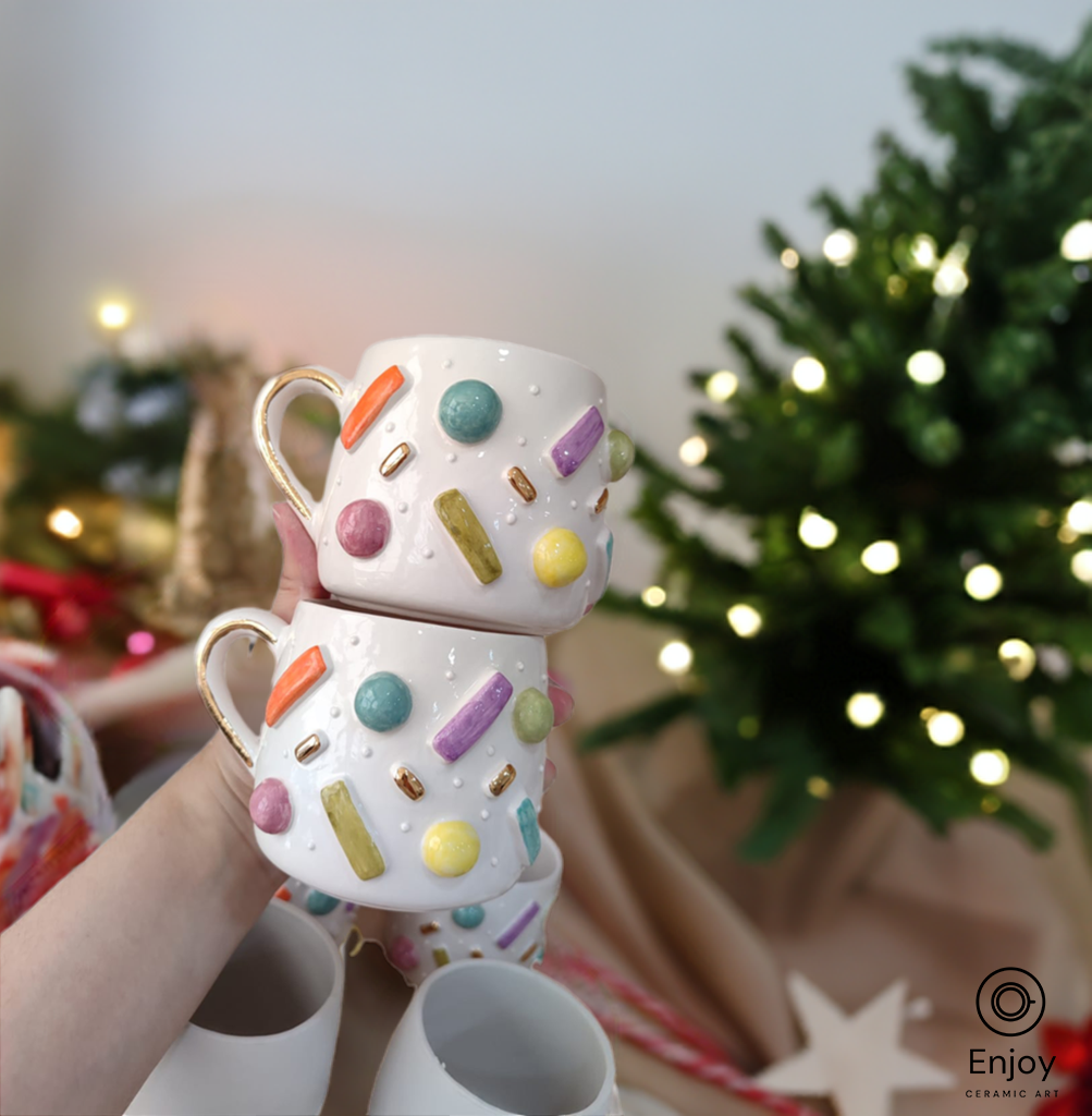 Hand holding two stacked ceramic mugs with colorful sprinkles design, set against a festive Christmas backdrop.