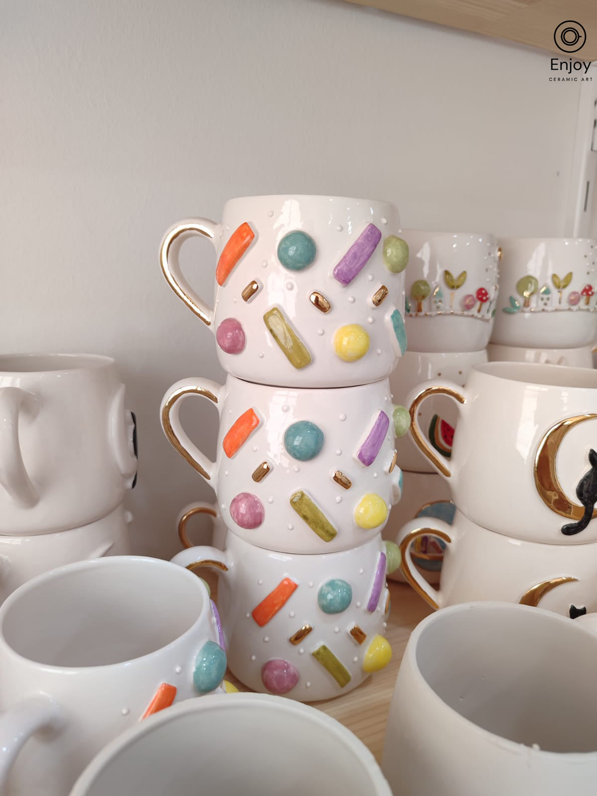 Three handmade mugs stacked on a wooden shelf, featuring vibrant pastel sprinkles in orange, yellow, green, purple, and pink with gold accents, offering a playful and cheerful aesthetic.