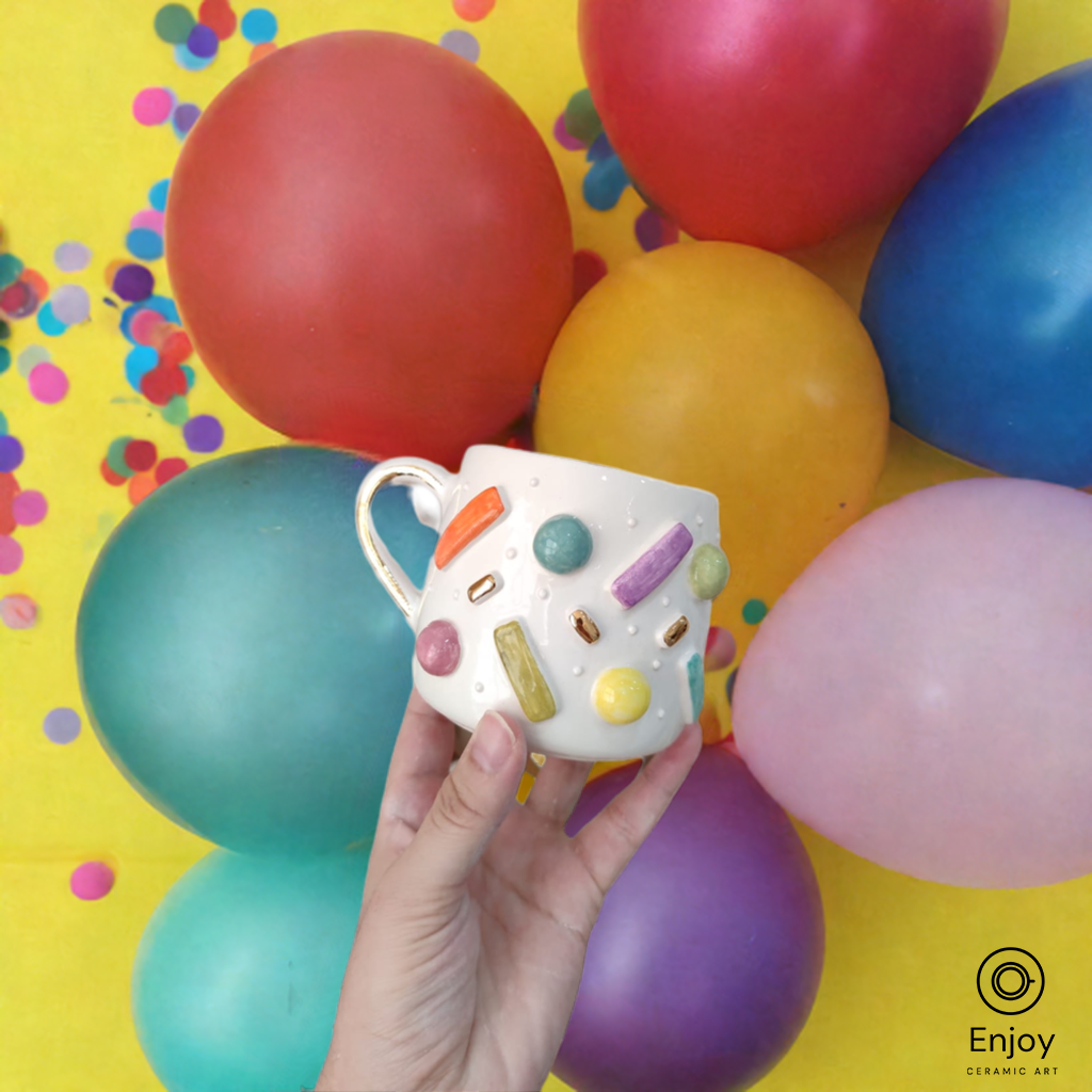 Hand holding a festive ceramic mug with colorful sprinkles, set against a backdrop of vibrant balloons and confetti.