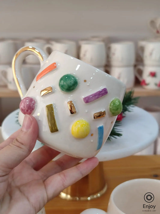 A vibrant handmade ceramic mug featuring colorful 'sprinkle' designs in various shapes and gold accents, with a gold handle, perfect for adding a playful touch to your coffee moments.