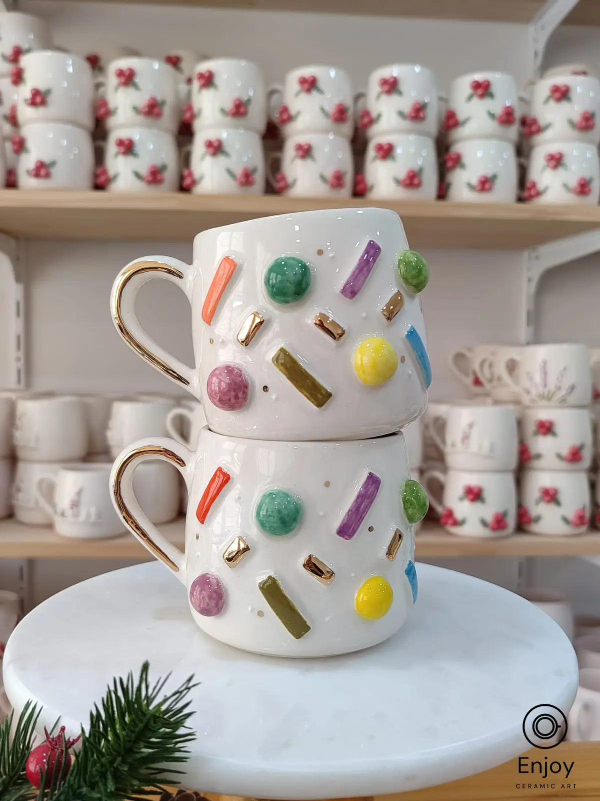 Handmade ceramic mugs with colorful candy-like sprinkles in various shapes, adorned with gold accents and a gold handle, stacked on a marble stand with festive greenery.