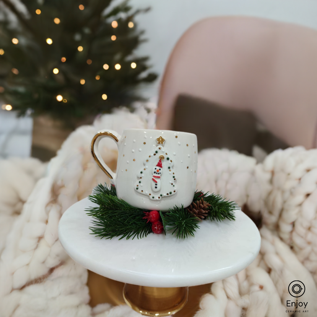 A cozy white mug with a 3D snowman and Christmas tree design, placed on a gold pedestal with festive greenery, in front of a softly lit Christmas tree.