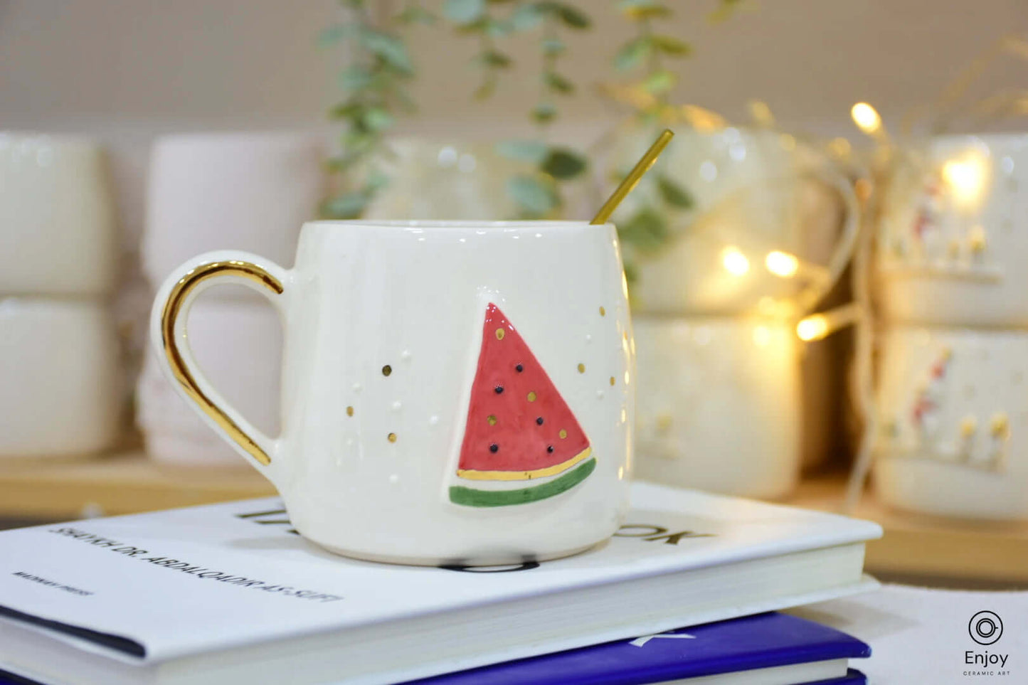 A handmade ceramic mug featuring a playful watermelon slice design with gold accents, placed on a book, perfect for a refreshing vibe.