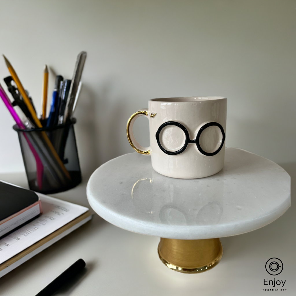 A whimsical ceramic mug featuring black glasses and a gold handle, set on a desk next to stationery, evoking a magical, studious vibe.